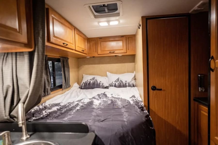 Cozy RV rental interior features mountain-themed bedding, overhead cabinets, and a compact kitchen area with a sink.