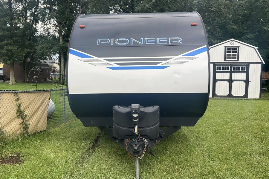 Front of a white and dark gray Pioneer travel trailer, ideal for rv rental. Parked on grass with sheds & trees visible.