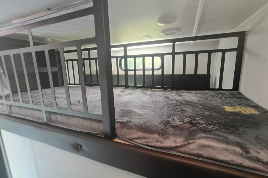 Top bunk bed in an RV rental, featuring a soft blanket and safety railing. A window is visible in the background.