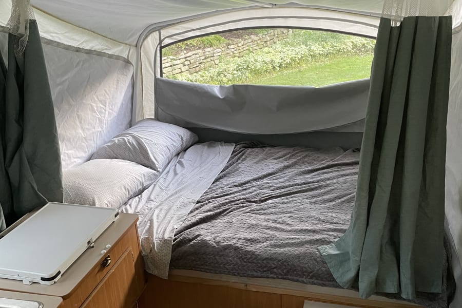 Interior of an RV rental pop-up camper featuring a bed with bedding, compact kitchen, and a window view of greenery.