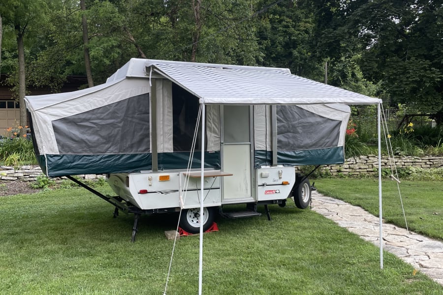 A Coleman pop-up camper is set up on grass with its awning extended, perfect for an RV rental getaway.