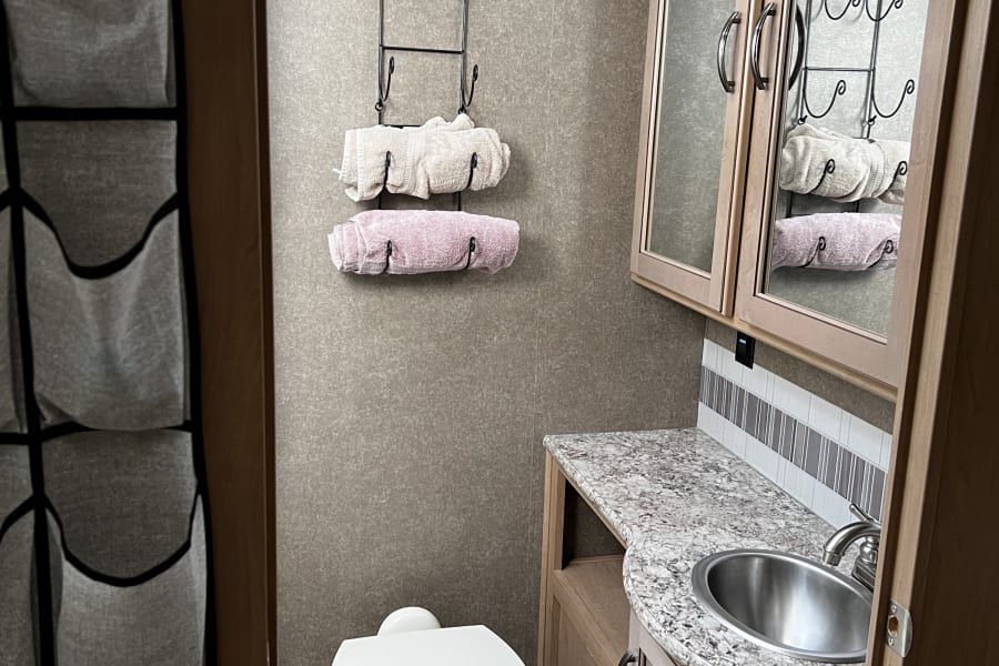 RV rental bathroom features a compact toilet, sink, mirrored cabinet, and wall-mounted towels, with hanging storage on the left.