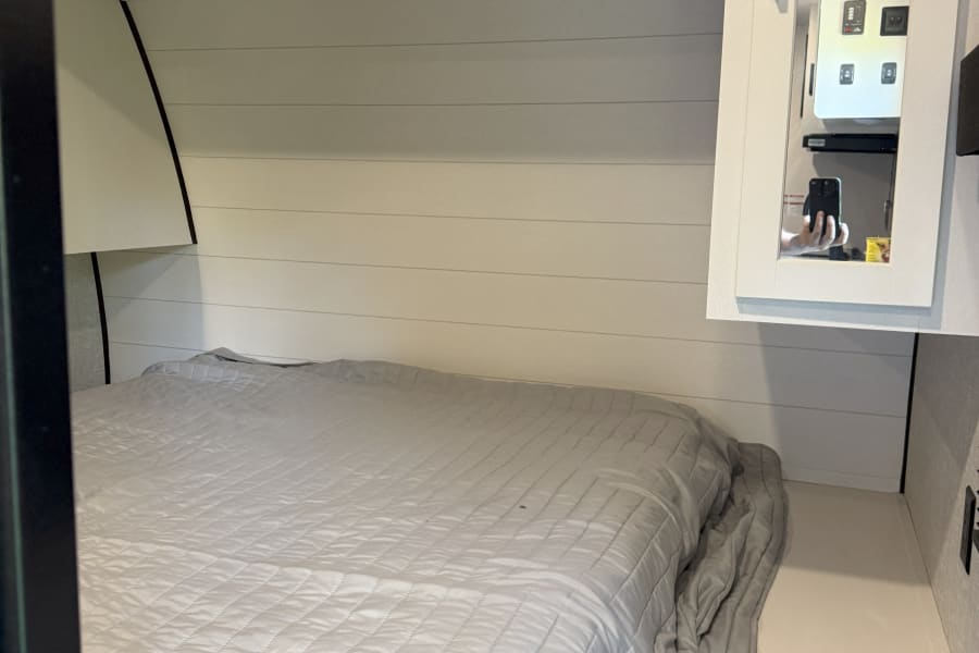 Compact RV rental bedroom featuring a light gray quilted bed, shiplap walls, and reflective cabinet.