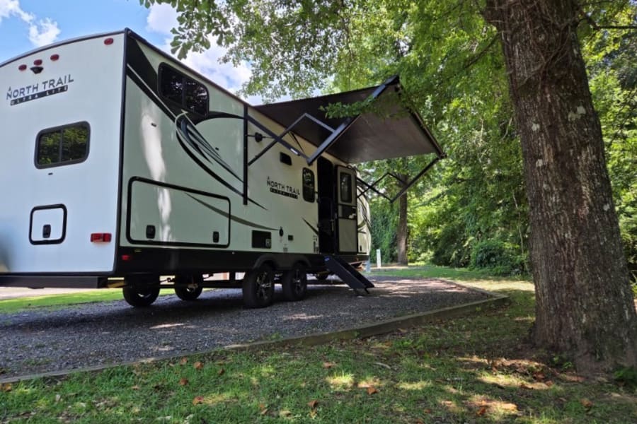 A North Trail RV rental with an extended awning is parked on a gravel campsite amidst lush trees, perfect for a getaway.