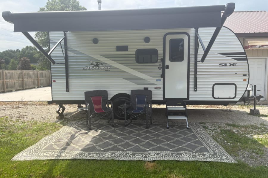 Side view of a white Jay Flight SLX RV rental, parked with an extended black awning. Two camping chairs and an outdoor rug are set up.