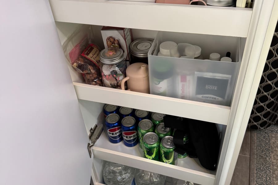 Inside an RV rental, a compact pantry unit neatly holds TAZO tea, snacks, Pepsi, water, and various travel essentials on shelves.