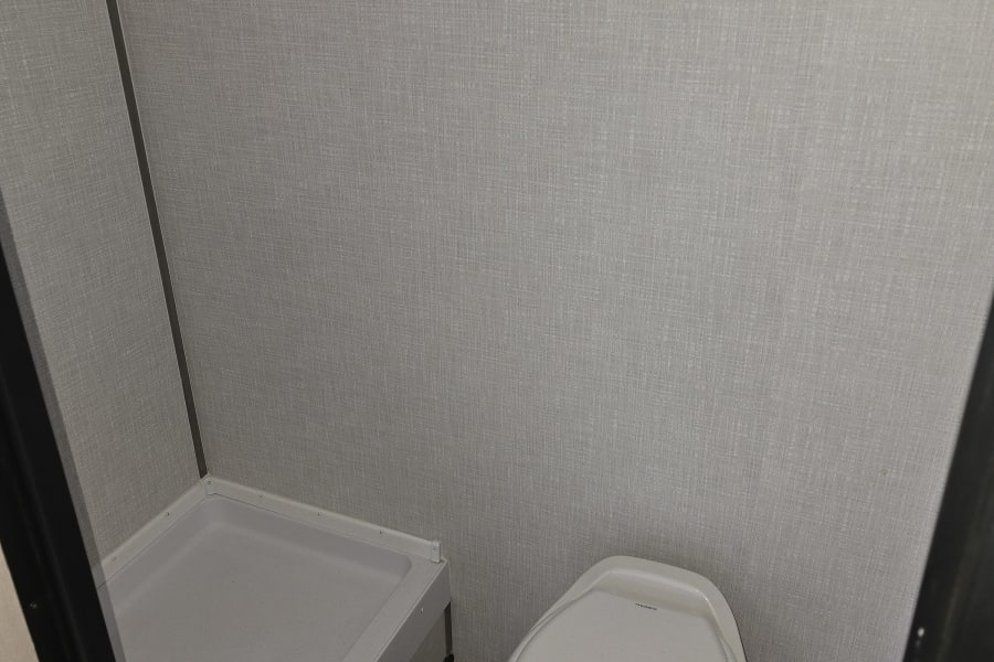 Inside an RV rental bathroom, featuring a compact shower and toilet. Walls are light gray, floor is wood-look.