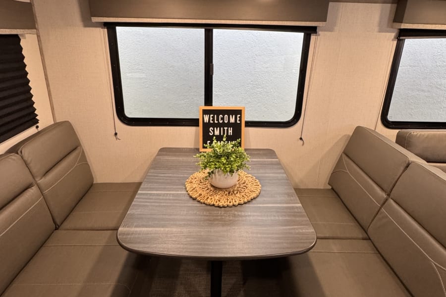 Cozy RV rental interior features a dinette with bench seats, table, 'Welcome Smith' sign and plant.
