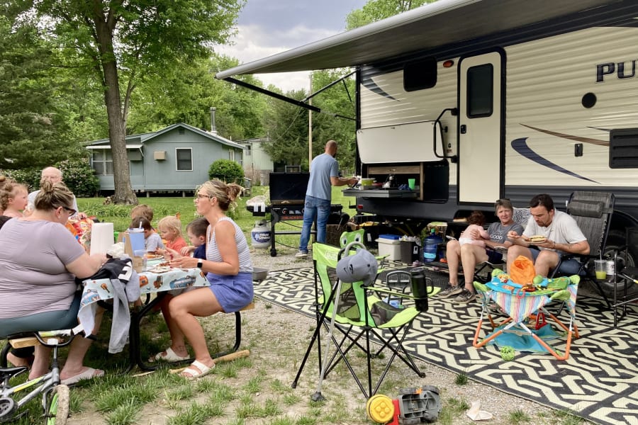 Family and friends enjoy an outdoor meal and grilling by their RV rental, with children and adults relaxing at the campsite.