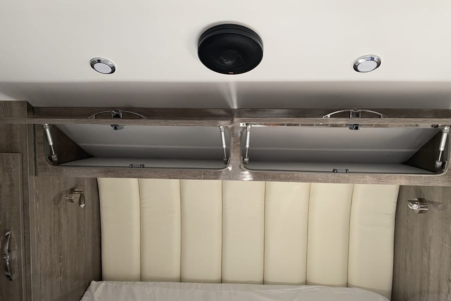 Comfortable RV rental bedroom with padded headboard, open overhead storage cabinets, and ceiling-mounted speaker/lights.