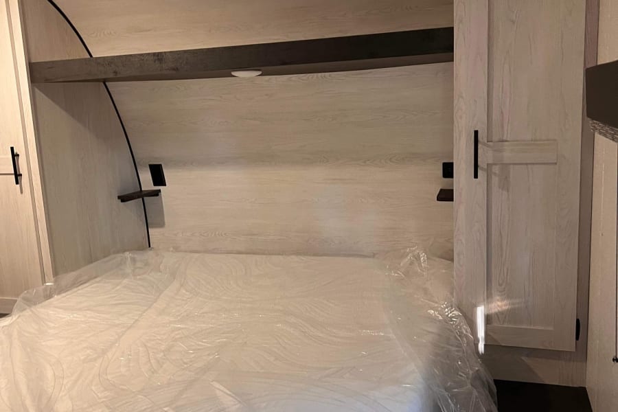 Compact RV rental bedroom featuring a new mattress still in plastic, with integrated storage and shelving units.