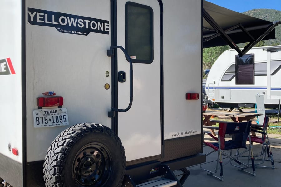 A white Yellowstone travel trailer, perfect for an rv rental, is parked at a sunny campsite with its awning out and outdoor seating.