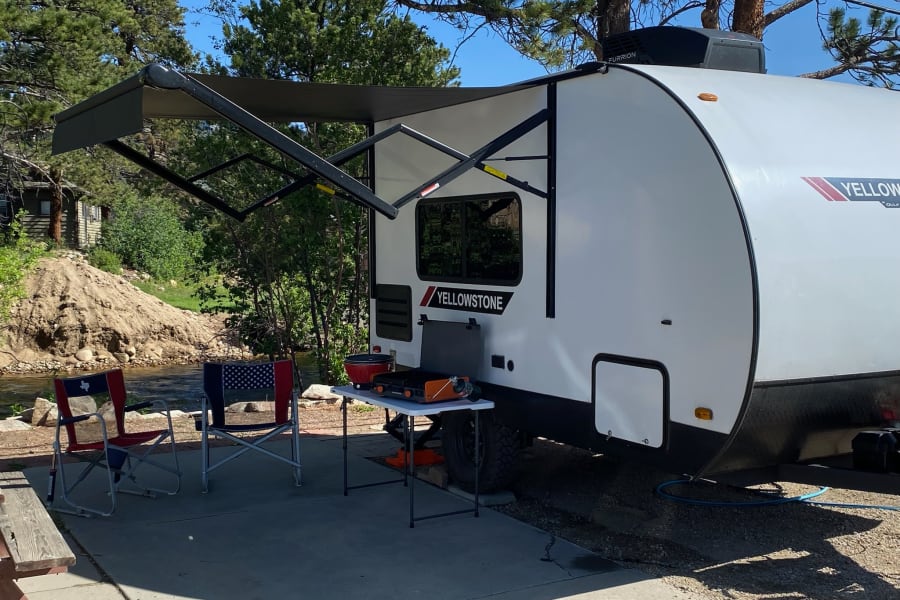 This Yellowstone rv rental provides a complete outdoor setup: extended awning, cooking station, and chairs by a scenic river campsite.