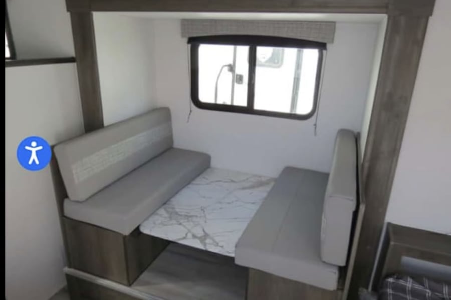 Inside an RV rental: a bright, modern dinette with two grey benches and a white marble-look table by a window.