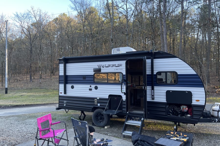 Family camping in a Wolf Pup **rv rental** travel trailer, with outdoor chairs at a scenic wooded site.