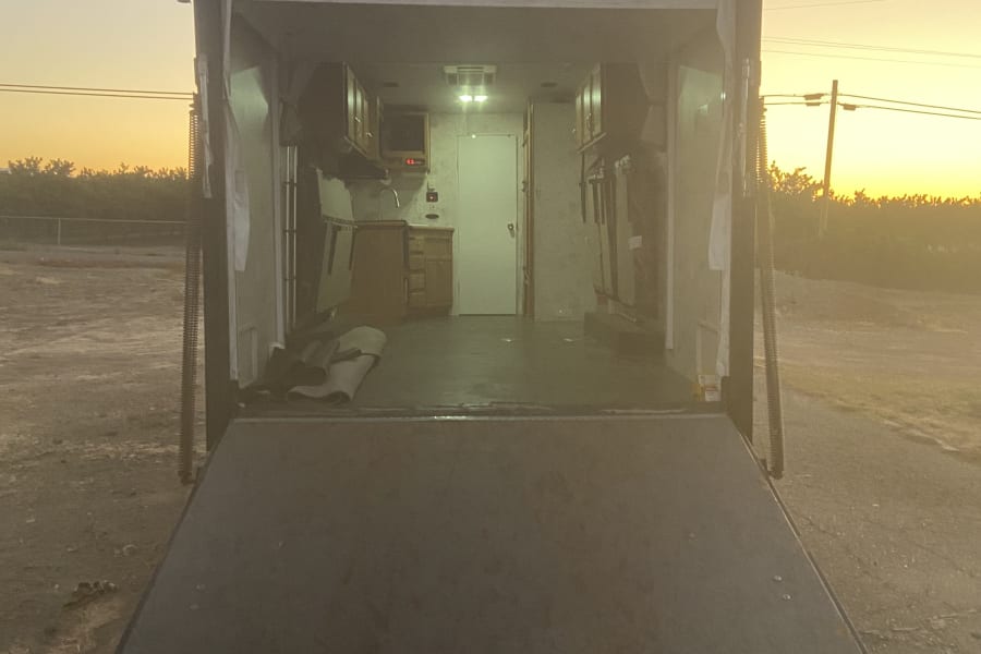 Inside an RV rental, the ramp door is open, showing a functional mobile living space with cabinets & a sink during sunset.