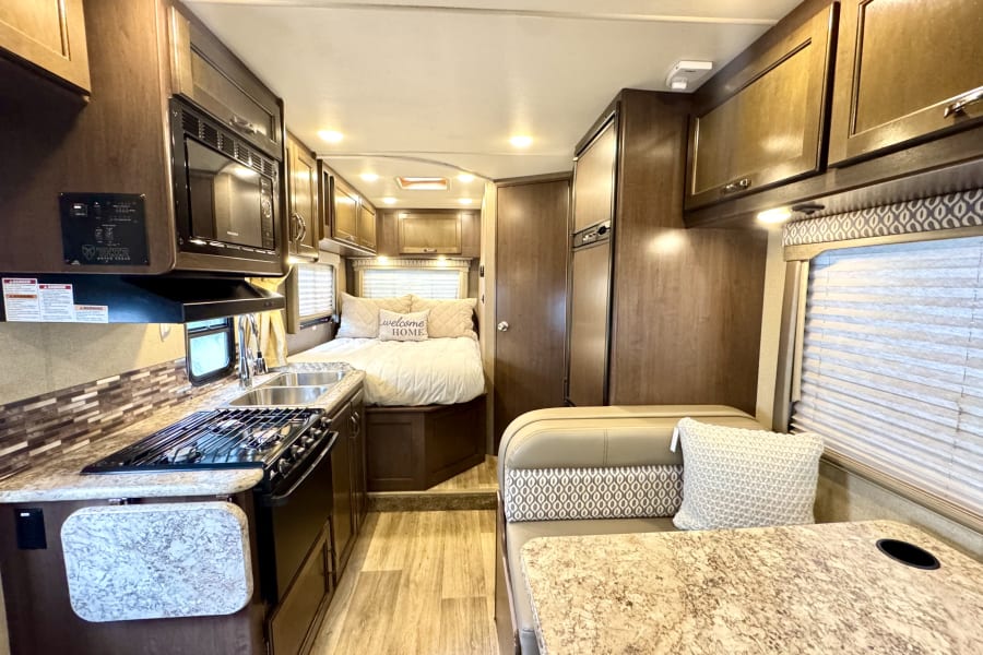 Interior of an RV rental: features kitchen with stove/sink, bed with "welcome HOME" pillow, and a dinette.