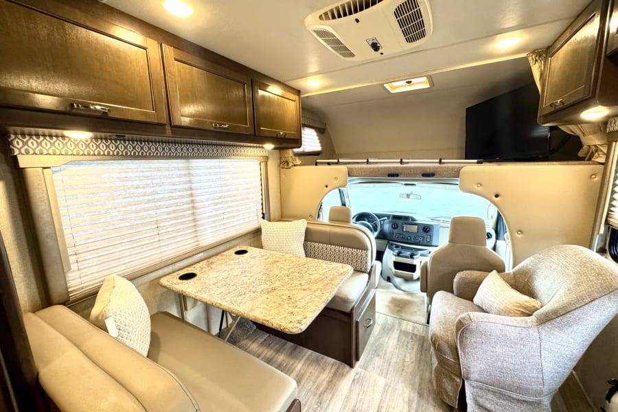 Spacious RV rental interior with dinette, lounge chair, overhead cabinets, and AC. Perfect for your road trip.
