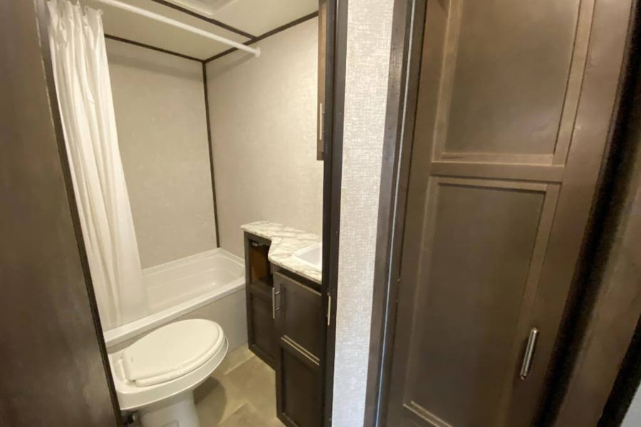 Inside an RV rental bathroom: a white toilet, compact tub/shower with curtain, and dark wood vanity with sink. Practical for travel.