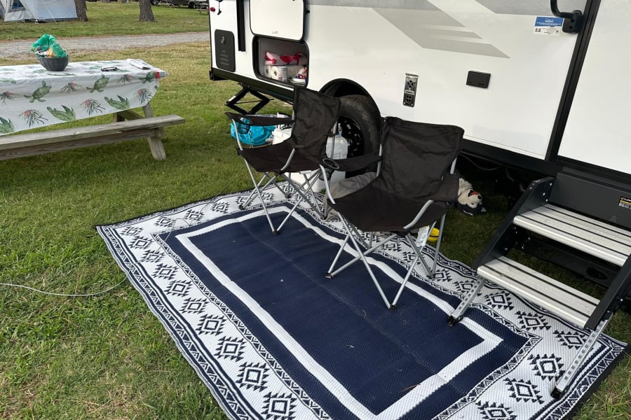 RV rental campsite setup: blue/white outdoor rug, two folding chairs, and picnic table on green grass for relaxation.