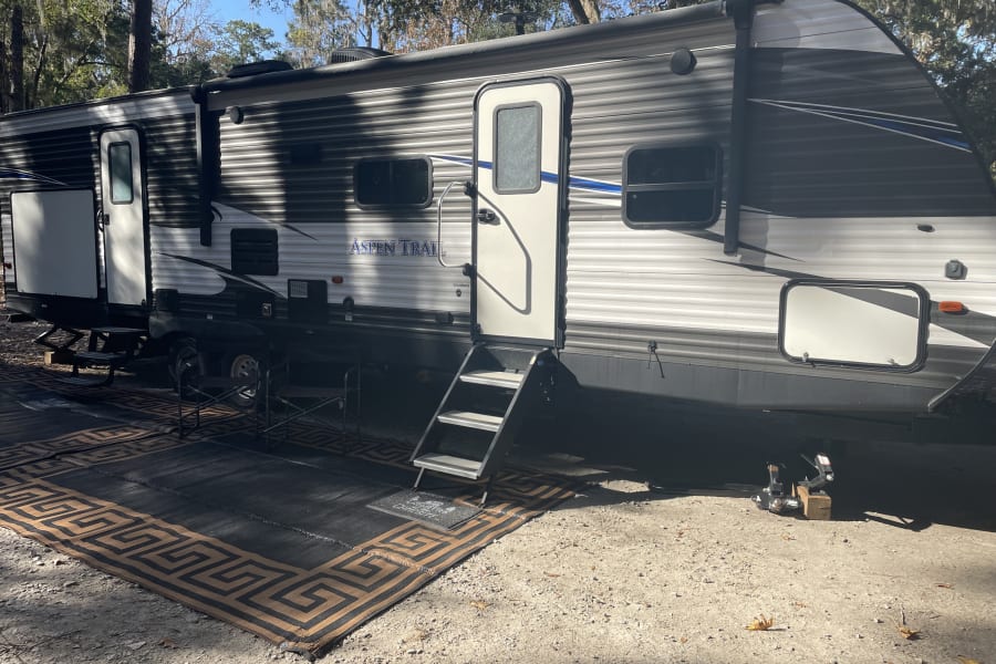 RV rental at a sunny campsite. An Aspen Trail travel trailer is set up with an outdoor mat and chairs.