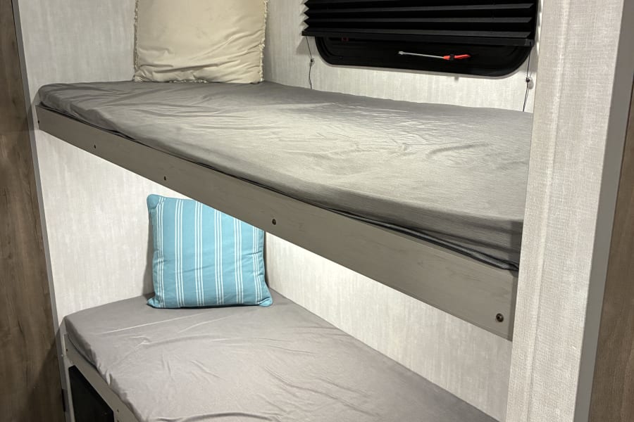 RV rental: Compact sleeping area with two bunk beds, fitted with grey sheets and pillows. Features window and under-bed storage.