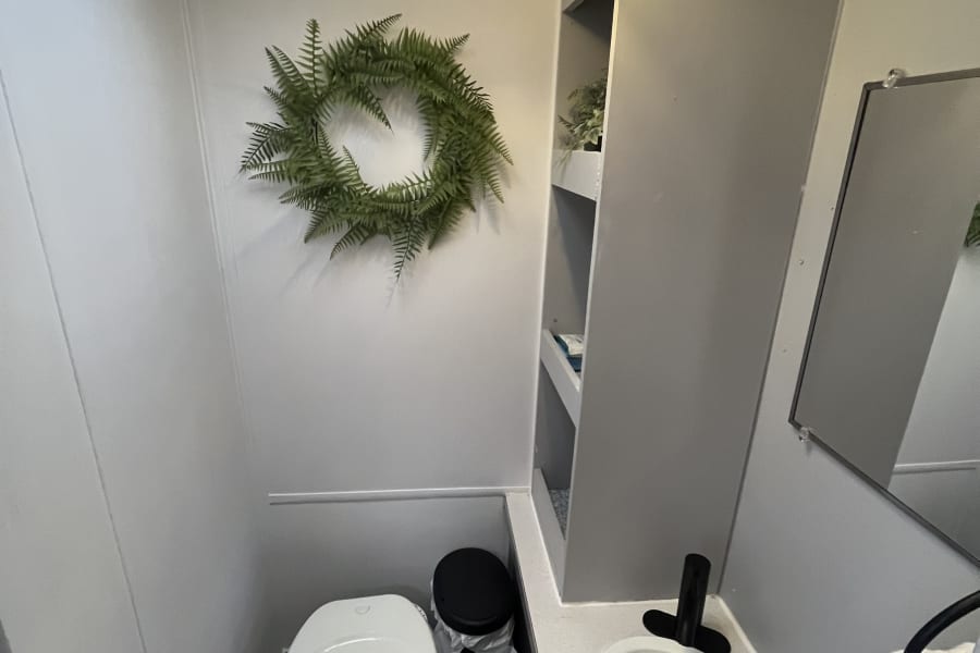Well-lit RV rental bathroom with compact toilet, sink, mirror, and storage shelves. Features modern grey and white decor.