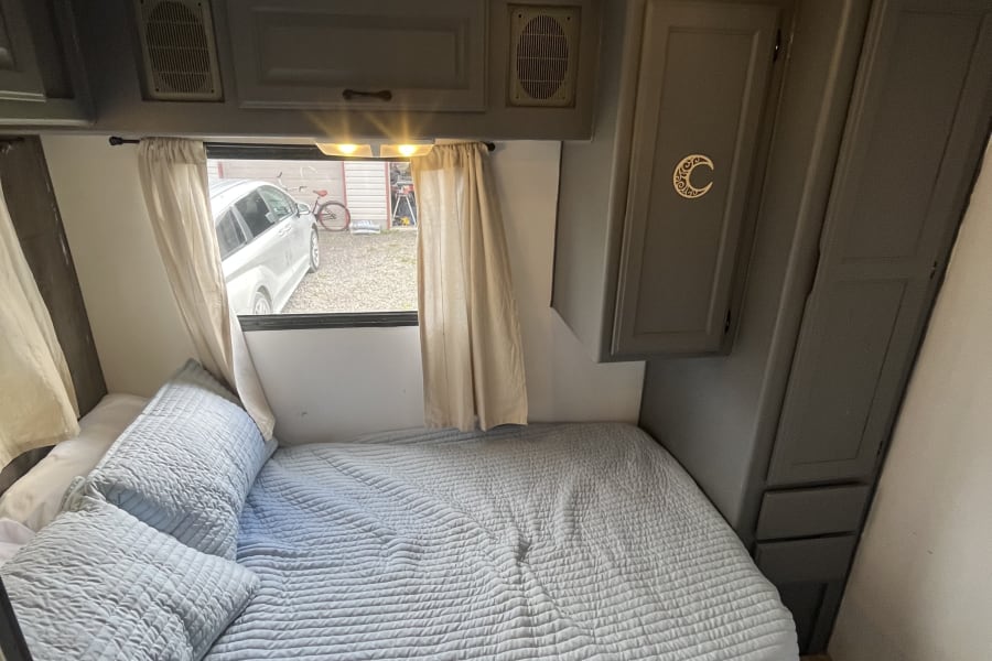 Cozy RV rental bedroom featuring a soft blue bed, window with curtains, and gray overhead cabinets.