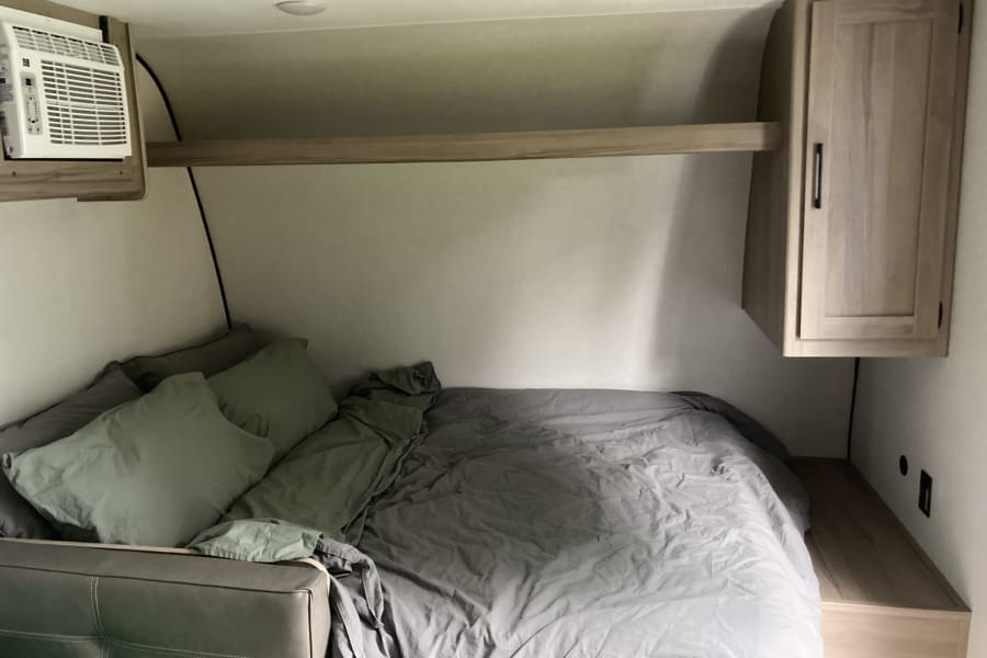 Cozy RV rental bedroom featuring a full bed with light bedding, a mounted AC unit, and an overhead wooden cabinet. Ideal for travel.