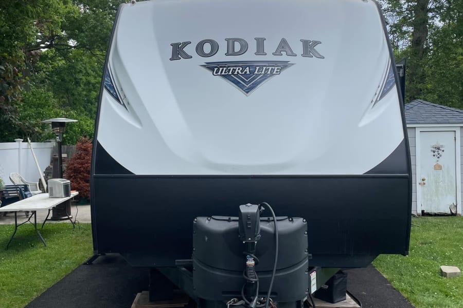 White Kodiak Ultra Lite RV rental trailer, parked on asphalt with trees and a shed visible in the background.