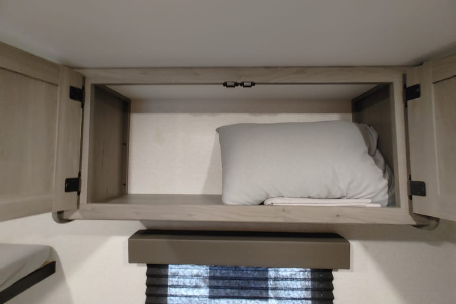 Overhead storage in an RV rental, showing a pillow and bedding in a light wood cabinet. Essential for travel comfort.