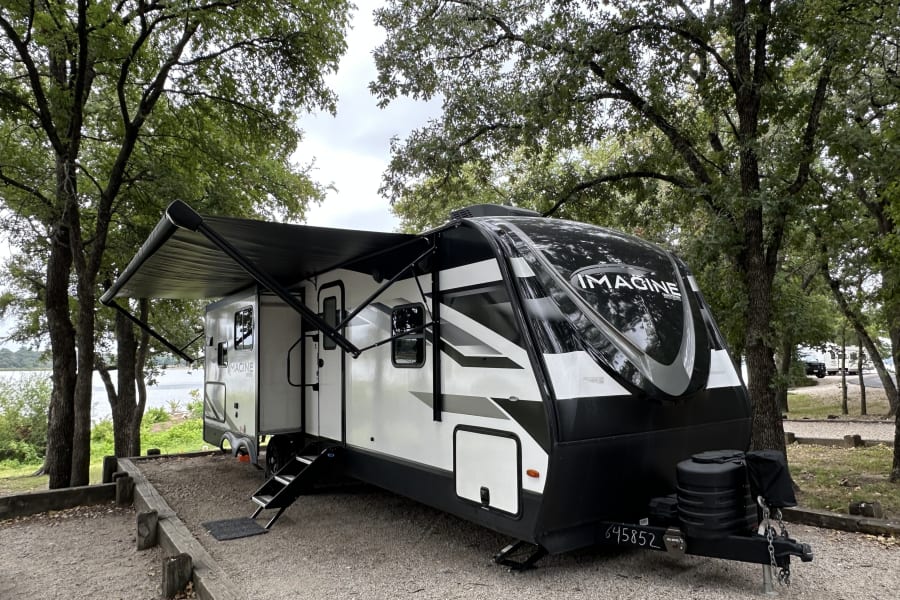 This RV rental is set up at a scenic lakeside campsite with its awning extended, surrounded by lush trees.