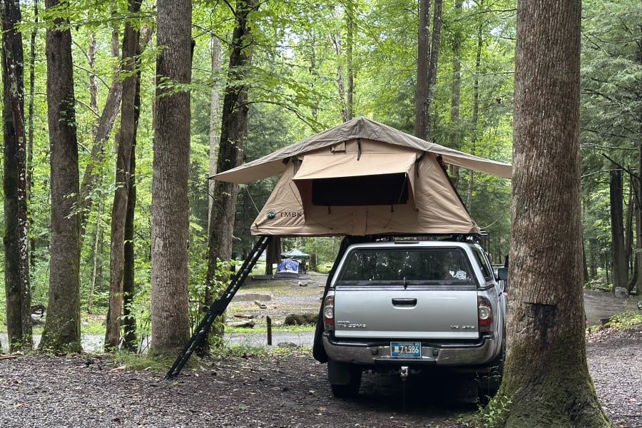 A silver pickup truck with a TMBK rooftop tent set up for camping in a lush forest campsite, an alternative to traditional RV rental.