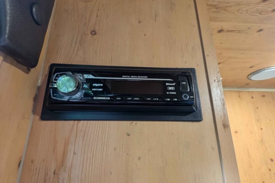 Inside an RV rental: a wooden wall with an Axerra car stereo installed. A white cable runs through a grommet above it.
