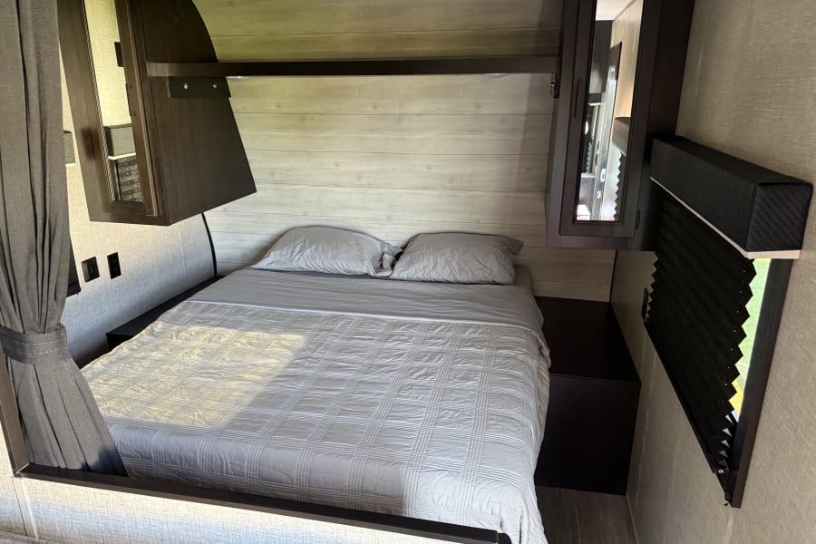 Inside an RV rental: a neatly made bedroom with a light gray bed, overhead storage, and window with blinds.