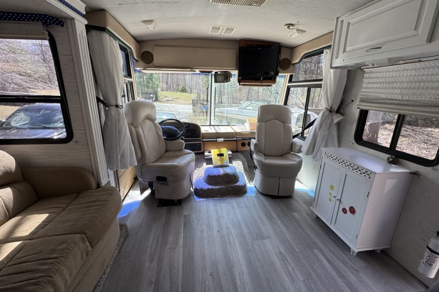 Interior of a bright RV rental: driver's cockpit, passenger seat, sofa, and modern gray flooring.