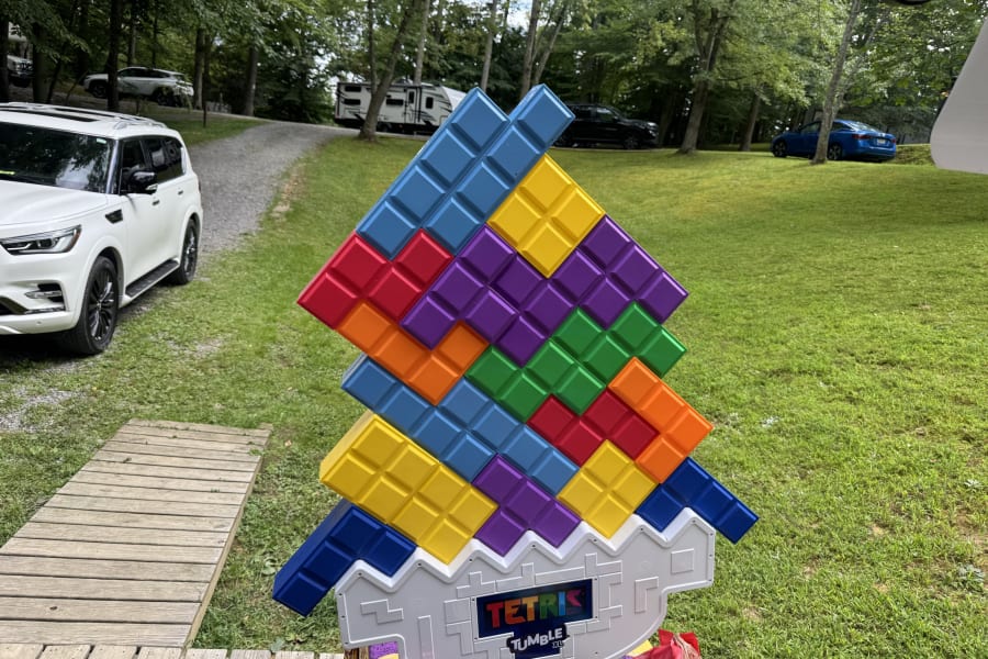 A giant Tetris Tumble game on grass at a campground, with an RV rental and other vehicles visible under trees.
