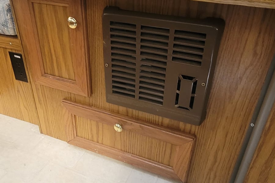 Inside an RV rental: close-up of wood cabinetry, a furnace vent, and light-colored flooring.
