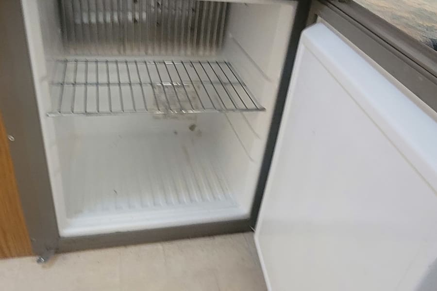 Inside an RV rental, a compact refrigerator is open, revealing empty wire shelves. Ready for your snacks!