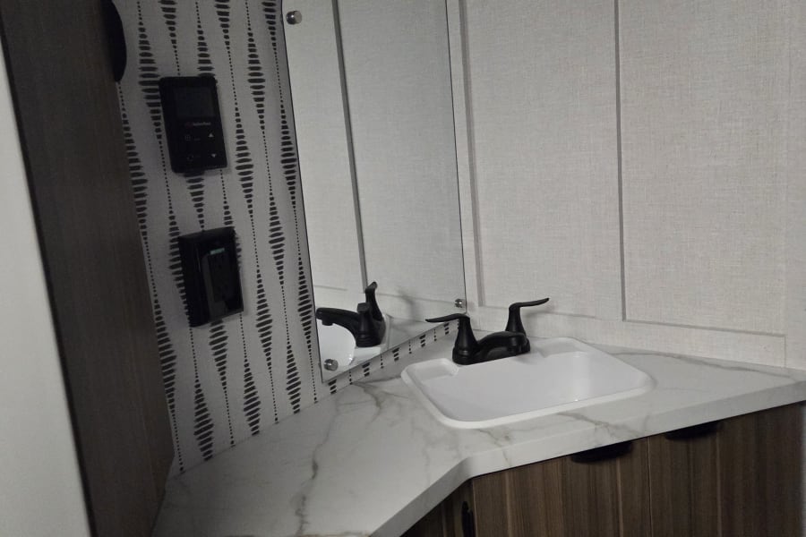 RV rental bathroom: modern vanity with white sink, black faucet, curved mirror, and patterned wallpaper. Includes paneling & controls.