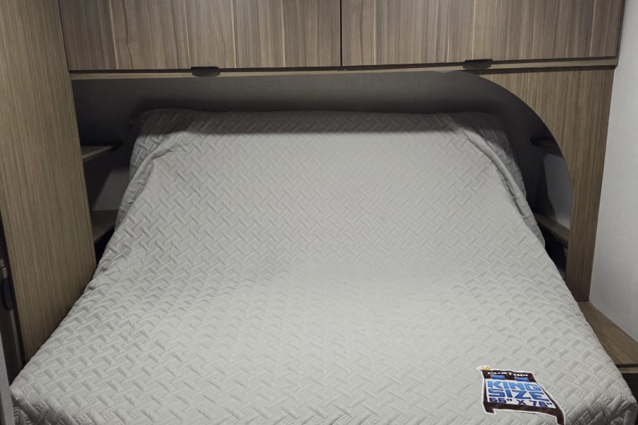 Brightly lit RV rental bedroom featuring a quilted custom king-size bed (66"x78") and integrated overhead storage.