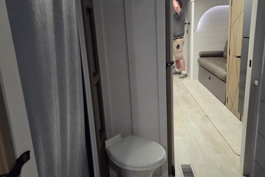 RV rental interior: compact bathroom with toilet and shower, opening to a bright living area with a sofa.