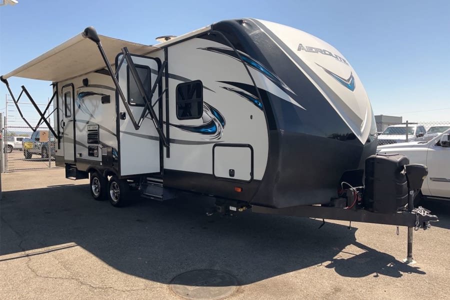 Explore the outdoors with this travel trailer RV rental. It has a modern white, black, and blue design with an extended awning.
