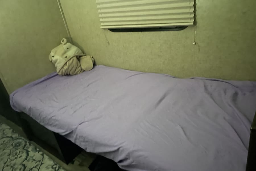 Cozy single bed inside an RV rental, complete with a plush toy, purple sheet, and window blinds. Ready for rest!