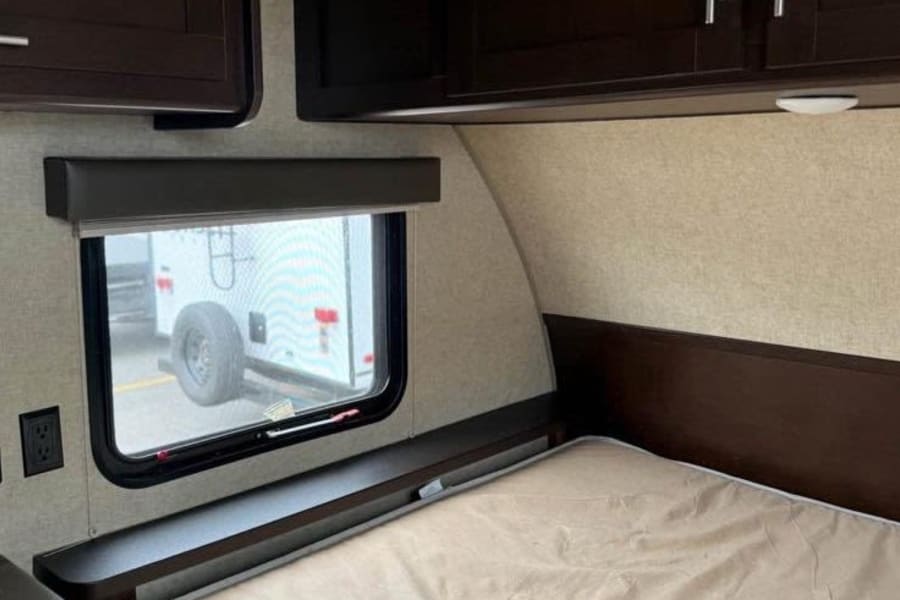 RV rental interior with bed, dark cabinets, window, and wall-mounted shelf. Ideal for a camper or travel trailer.