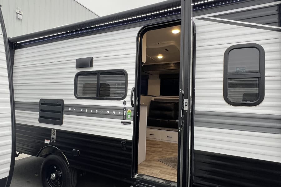 This black and white **RV rental** has its door open, with steps leading to a visible interior (bunk beds). An awning with LED lights is extended.