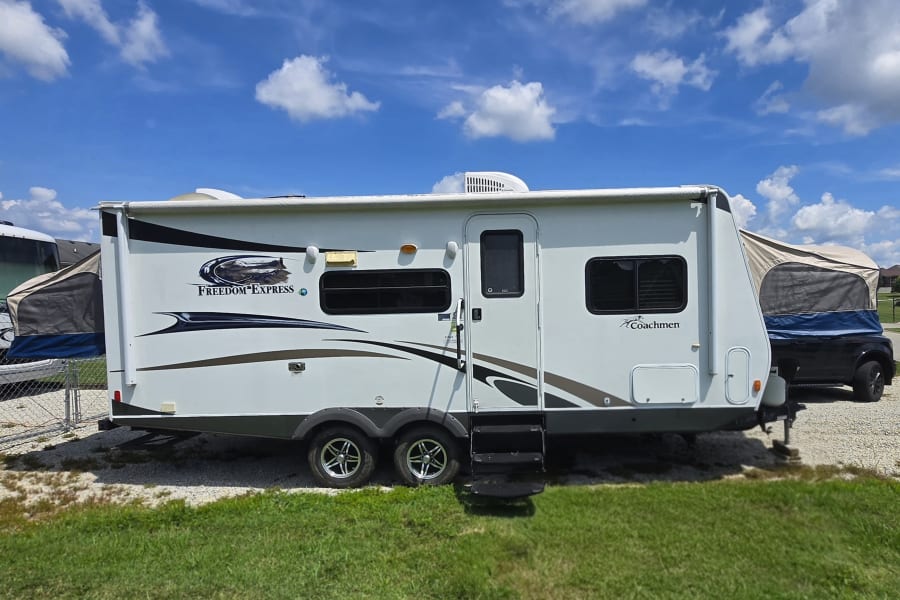 White Coachmen Freedom Express travel trailer RV rental, parked on gravel under a blue sky.