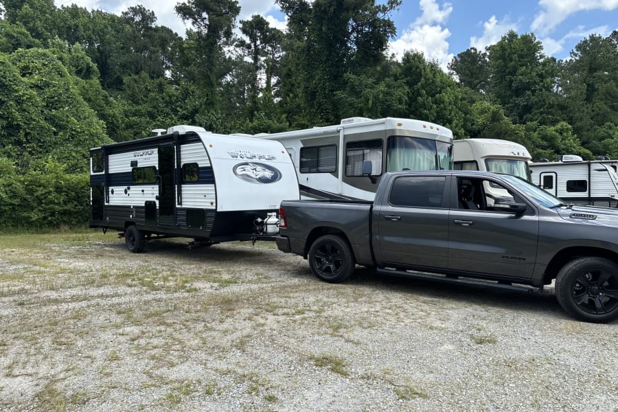 A gray pickup truck is hitched to a travel trailer on a gravel lot, with other RVs and trees in the background. Ideal for RV rental.