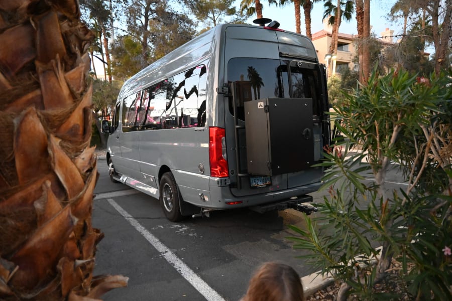 A gray Mercedes-Benz Sprinter 'Passage' RV rental, equipped with a rear storage box, is parked among palm trees.
