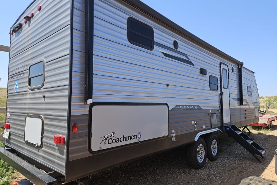 A large, modern Coachmen travel trailer, parked outdoors on gravel. Consider this RV rental for your next trip!